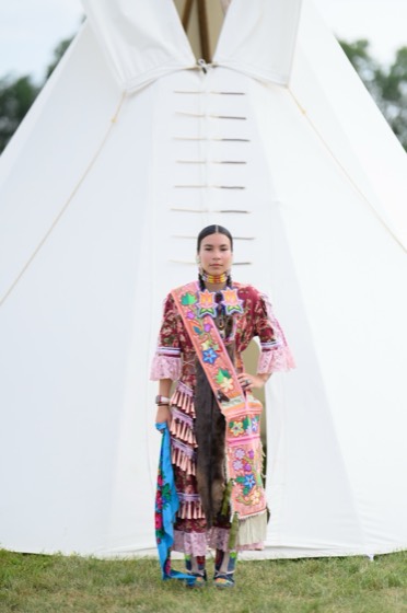 Valerie Leith standing in full jingle dress regalia in front of a tipi