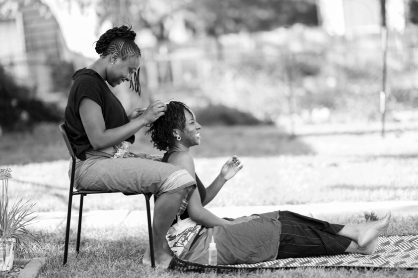 Deino and Gomoson Wonosikou in the yard, one on a chair braiding the other's hair as she rests on a mat in the grass