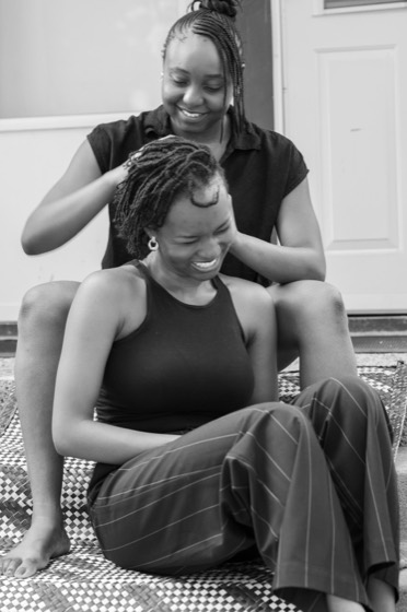 Deino and Gomoson Wonosikou on the front steps, one braiding the other's hair as she sits on a woven mat
