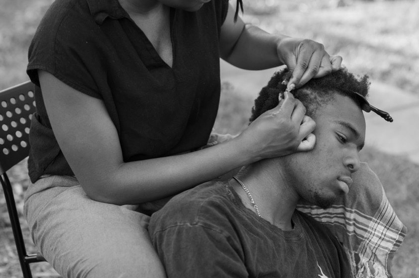 Gideon Wonosikou resting his head against Deino's knee while she parts a section of his hair at the temple