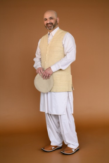 Wajih Mashadi standing in white salwar kameez and cream waistcoat, holding a dhol