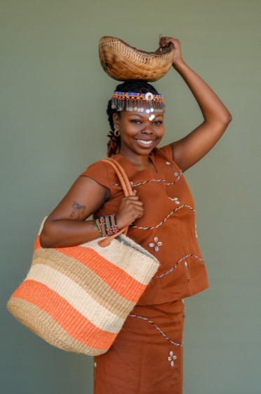 Lizzy Nyoike in Kikuyu regalia, a woven basket at her shoulder and a gourd balanced on her head