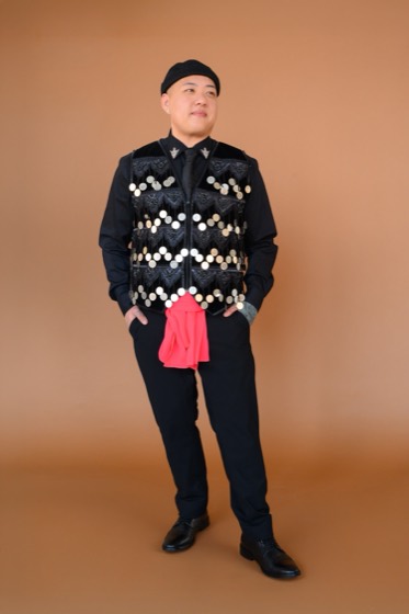 Remmus Xiong standing in a black Hmong vest hung with silver coins, pink sash at his waist